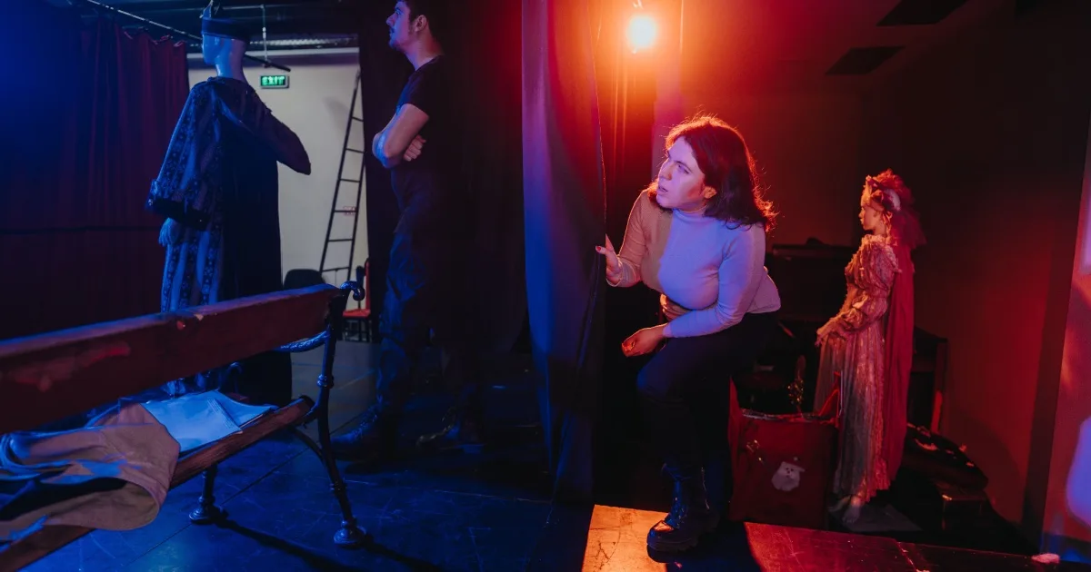 Theatrical performance scene with actors on stage under blue and red lighting, including a woman in center stage gesturing expressively.