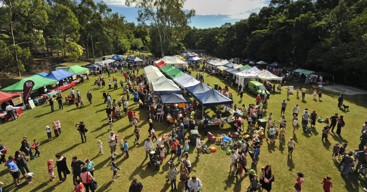 Large community outdoor festival with colorful tents and stalls on a green lawn surrounded by trees, with many attendees walking around.