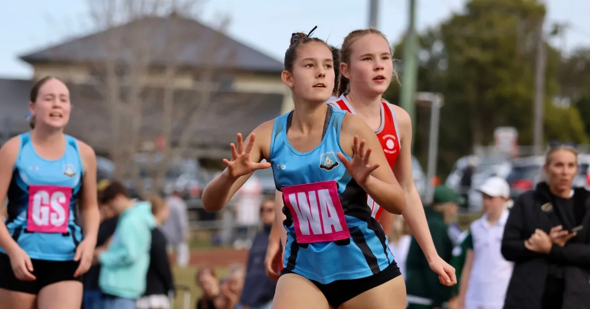 Young female netball players running during a match, wearing blue and red uniforms with "GS" and "WA" bibs at an outdoor sports venue.