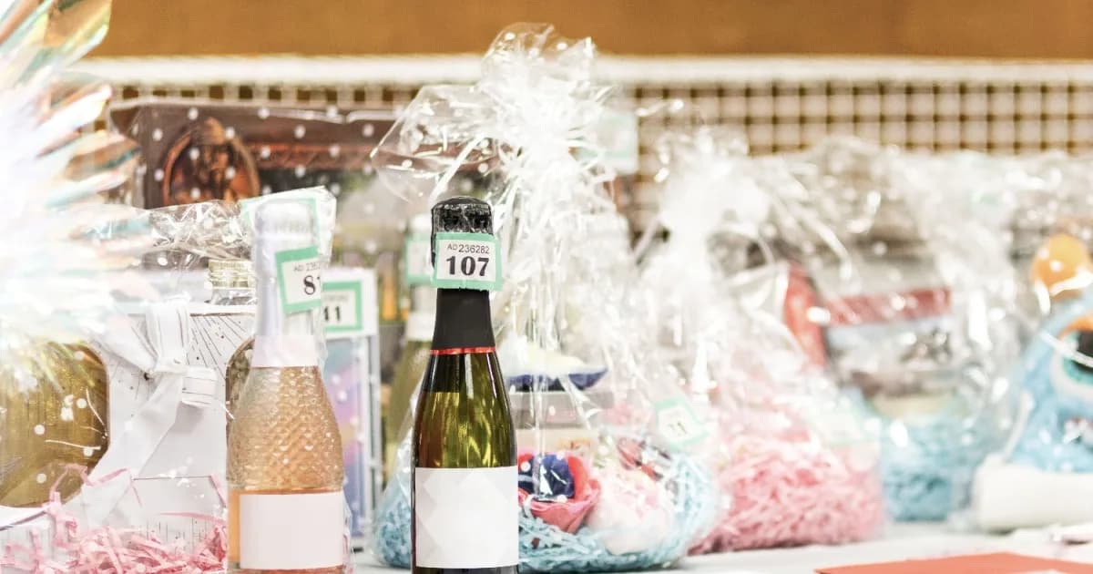 Auction item 107: bottle of champagne wrapped in cellophane with white bow, surrounded by gift baskets and wrapped items.