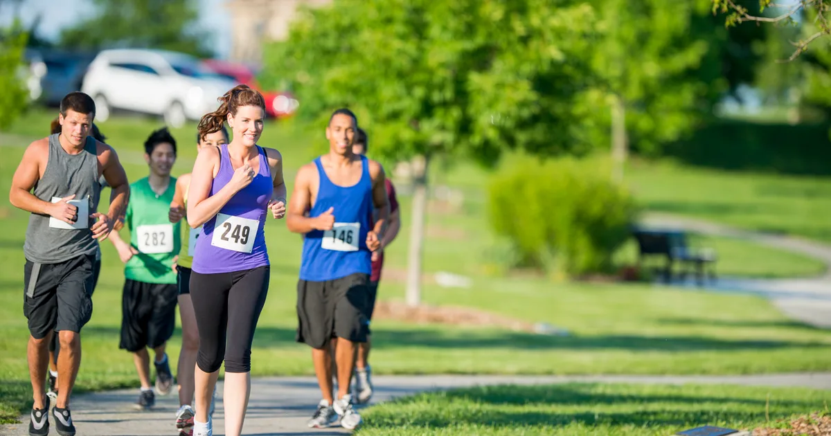 Group of runners participating in a community 5K race on a sunny day in a park, wearing race bibs with numbers.