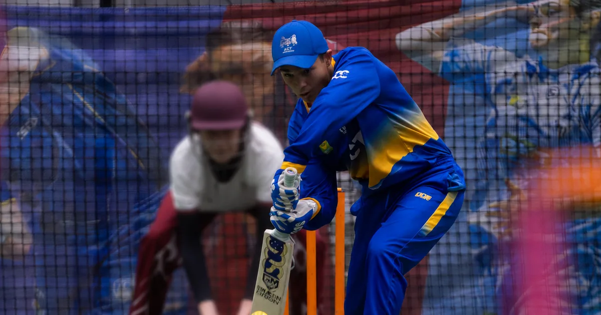 Cricket player in blue and gold uniform batting in nets during practice session with wickets visible.