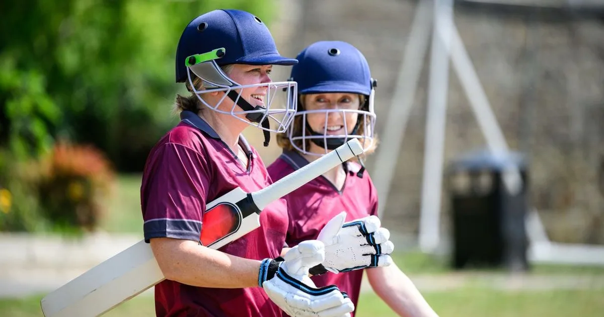 Two young female cricket players in maroon uniforms and blue helmets with protective gear, holding bats and smiling together.