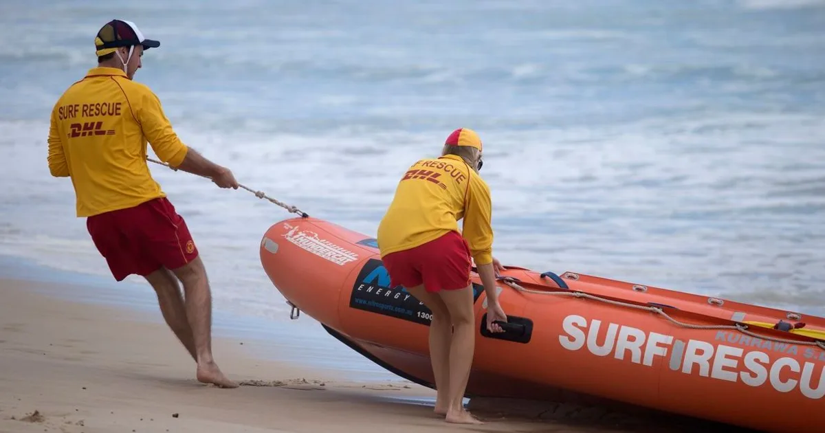 Two surf rescue lifeguards in yellow and red uniforms prepare an orange rescue boat on the beach.
