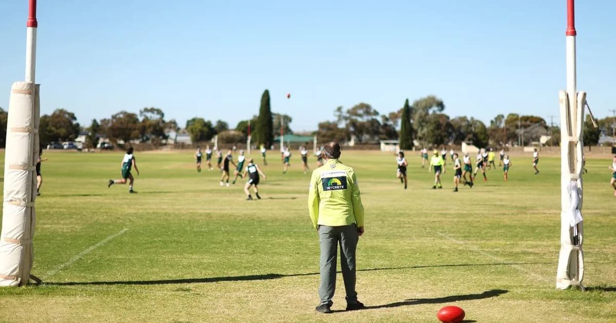 Goal umpire in yellow vest stands on Australian Rules Football field watching players during community football game.