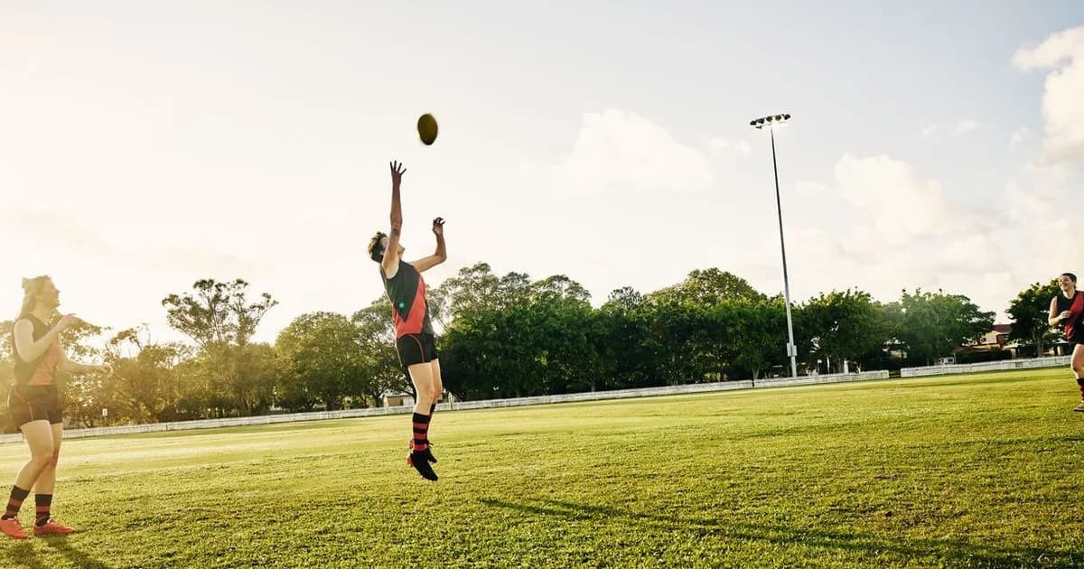 Australian Rules Football player jumps to mark the ball during a sunny afternoon game at an outdoor oval.