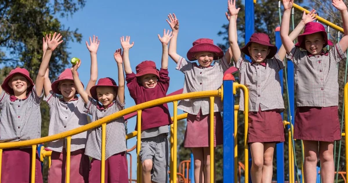 Happy school children in uniform and maroon hats cheering with raised arms on colorful playground equipment outdoors.