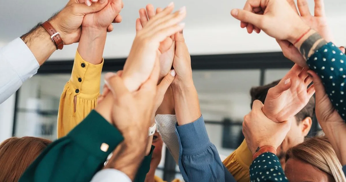 Group of diverse women joining hands together in a team huddle, celebrating unity and collaboration in a bright office setting.