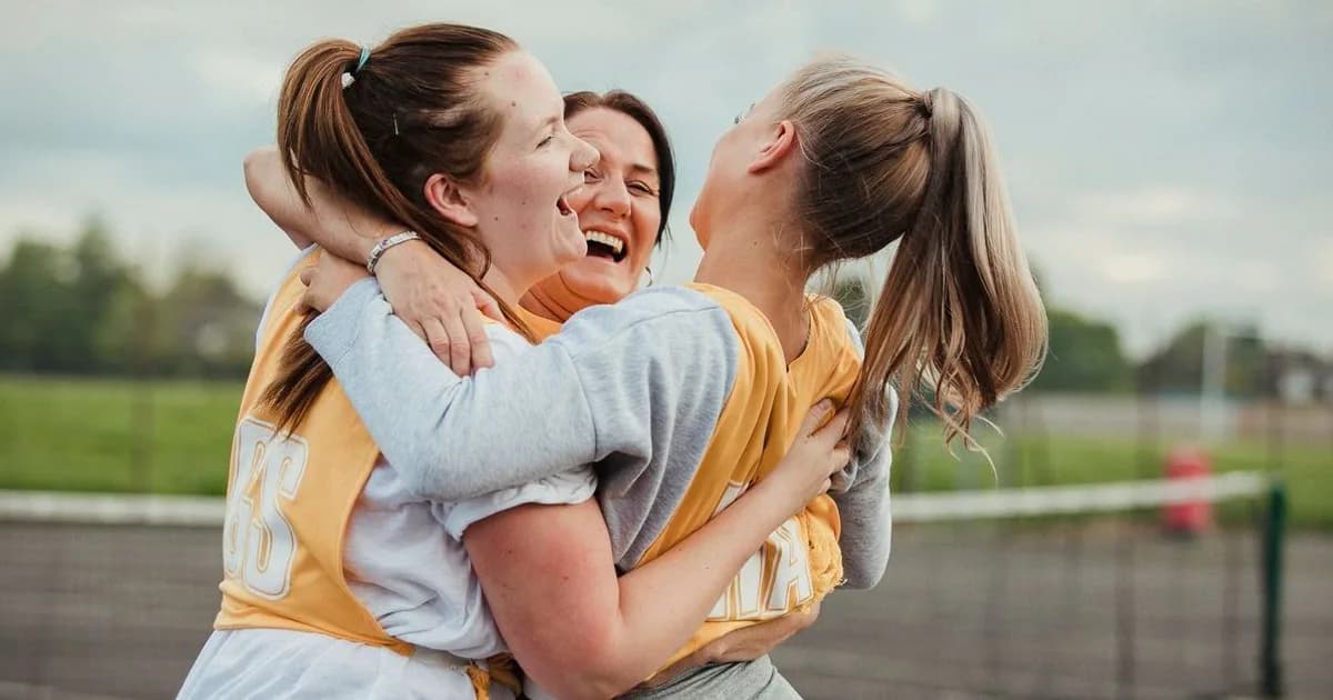 Three young female athletes in yellow sports uniforms embrace and laugh together outdoors by a field.