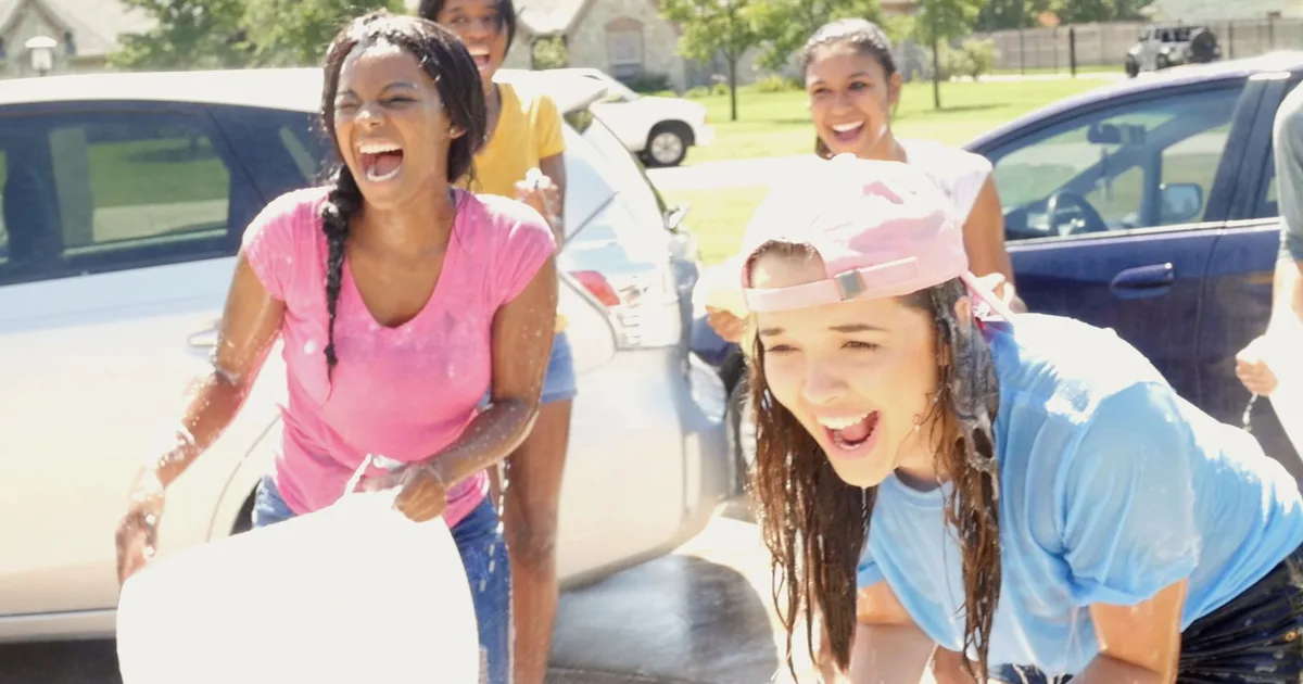 Girls having fun at a car wash fundraiser, spraying water and smiling while washing vehicles on a sunny day.