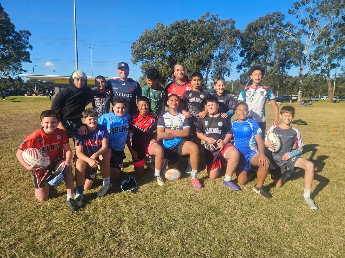 Youth rugby team and coaches pose together on a grass field on a sunny day, smiling at the camera.